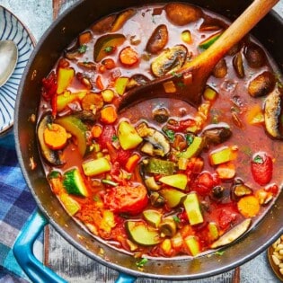 homemade vegetable soup in a dutch oven with a wooden spoon next to a bowl with two silver spoons.